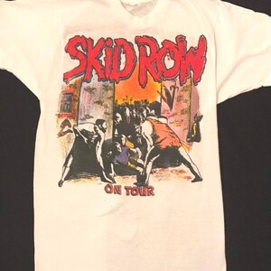 Op de afbeelding: Wit t-shirt met de bandnaam "SKID ROW" in rode en blauwe letters. Daaronder een afbeelding van mensen in een straatbeeld, met de woorden "ON TOUR" eronder gedrukt.