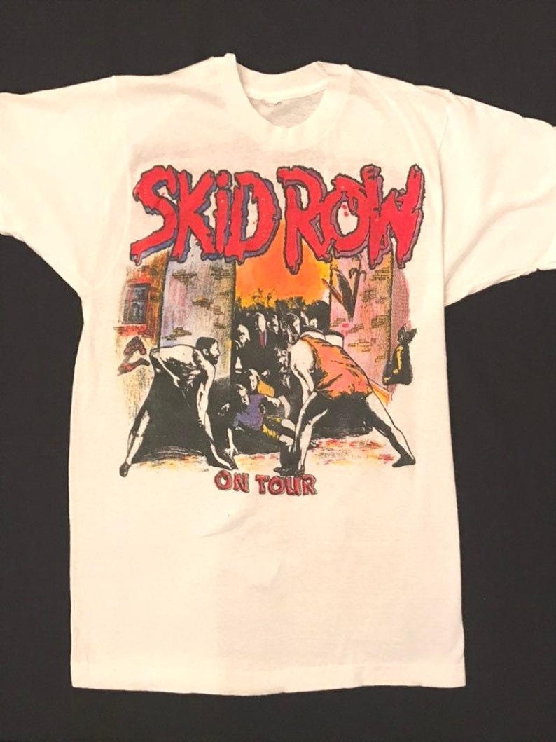 Op de afbeelding: Wit t-shirt met de bandnaam "SKID ROW" in rode en blauwe letters. Daaronder een afbeelding van mensen in een straatbeeld, met de woorden "ON TOUR" eronder gedrukt.