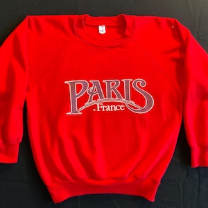 May include: A bright red sweatshirt with the word "PARIS" in a stylized font across the chest. Below the word "PARIS" is the word "France". The sweatshirt has long sleeves and a crew neck.