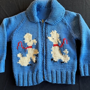 May include: A blue knitted cardigan with a zipper closure and two white poodle appliques on the front. The poodles are wearing red ribbons.