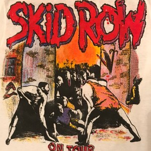 Op de afbeelding: Wit T-shirt met de bandnaam "SKID ROW" in rood, omlijnd met blauw, boven een afbeelding van een menigte. De woorden "ON TOUR" staan eronder in het rood.