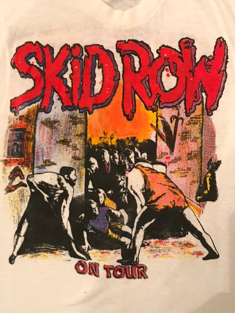 Op de afbeelding: Wit T-shirt met de bandnaam "SKID ROW" in rood, omlijnd met blauw, boven een afbeelding van een menigte. De woorden "ON TOUR" staan eronder in het rood.