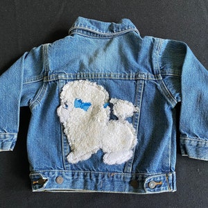 Puede incluir: Una chaqueta vaquera azul con una aplicación de perro blanco y esponjoso en la espalda.