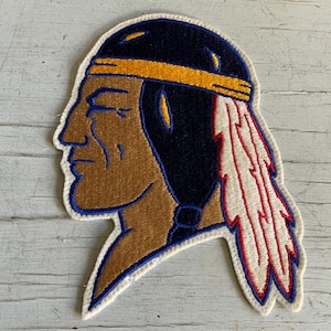 May include: Embroidered patch featuring a profile of a Native American man. The patch has a brown face, a navy blue headdress with a yellow band, and a red and white feather. The patch is outlined in blue and white.