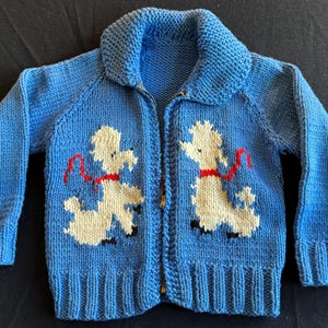 May include: A blue knitted cardigan with a zipper closure features two white poodle appliques with red collars. The poodles are facing opposite directions and are holding red leashes.