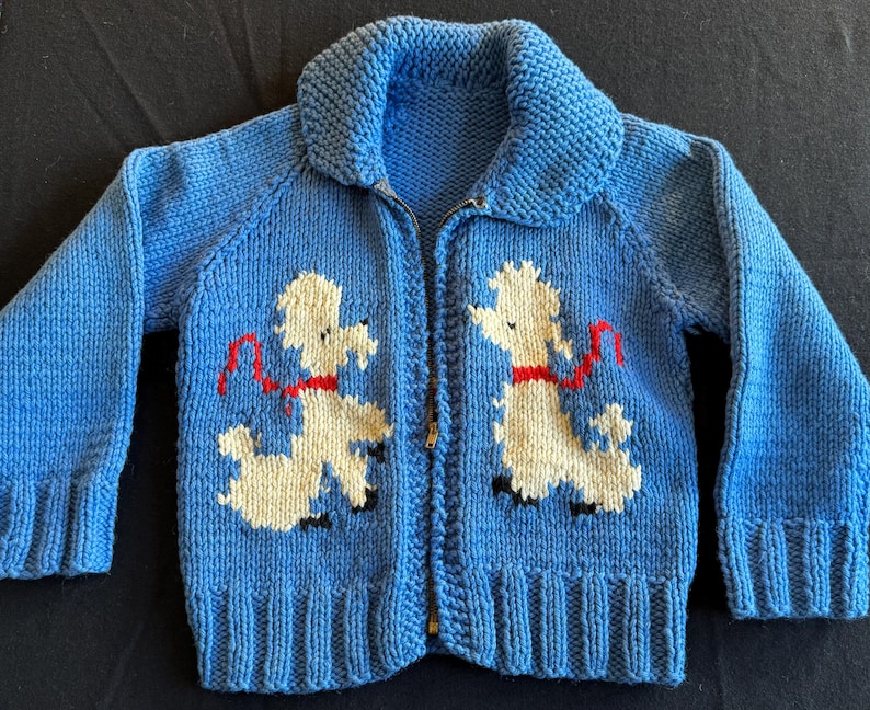 May include: A blue knitted cardigan with a zipper closure features two white poodle appliques with red collars. The poodles are facing opposite directions and are holding red leashes.