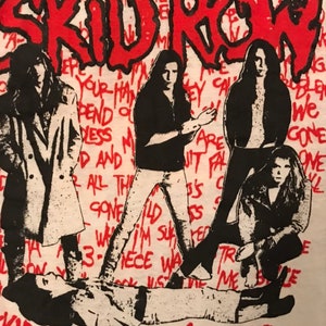 Op de afbeelding: Wit T-shirt met een afbeelding van de band Skid Row in concert. Het ontwerp bevat de naam van de band in het rood, bandleden in zwart en wit en rode tekst op de achtergrond.