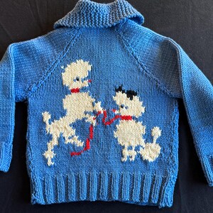 May include: A blue knitted sweater with a white poodle design. The poodle is holding a red string in its mouth.