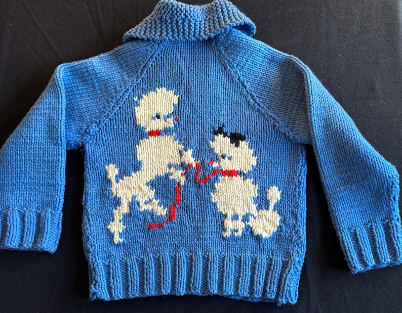 May include: A blue knitted sweater with a white poodle design. The poodle is holding a red string in its mouth.