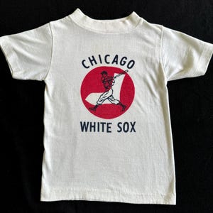 K&ouml;nnte beinhalten: Wei&szlig;es T-Shirt mit einem roten Kreis und einer Grafik eines Baseballspielers. Der Text "CHICAGO WHITE SOX" ist auf das Shirt gedruckt.