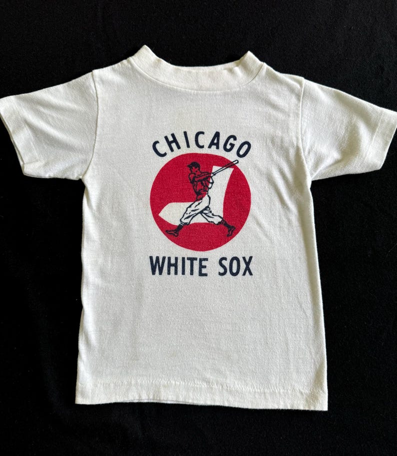 K&ouml;nnte beinhalten: Wei&szlig;es T-Shirt mit einem roten Kreis und einer Grafik eines Baseballspielers. Der Text "CHICAGO WHITE SOX" ist auf das Shirt gedruckt.