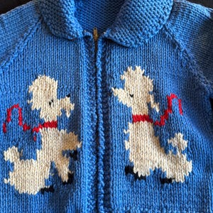 May include: A blue knitted cardigan with a zipper closure and a white poodle design on the front. The poodle is wearing a red ribbon.