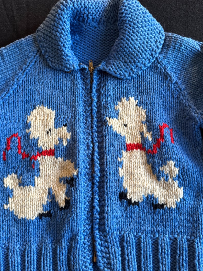 May include: A blue knitted cardigan with a zipper closure and a white poodle design on the front. The poodle is wearing a red ribbon.