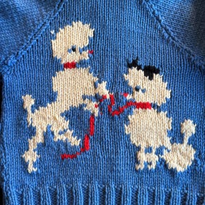 May include: A blue knitted sweater with a white and red poodle design. The sweater has a ribbed hem and a collar.