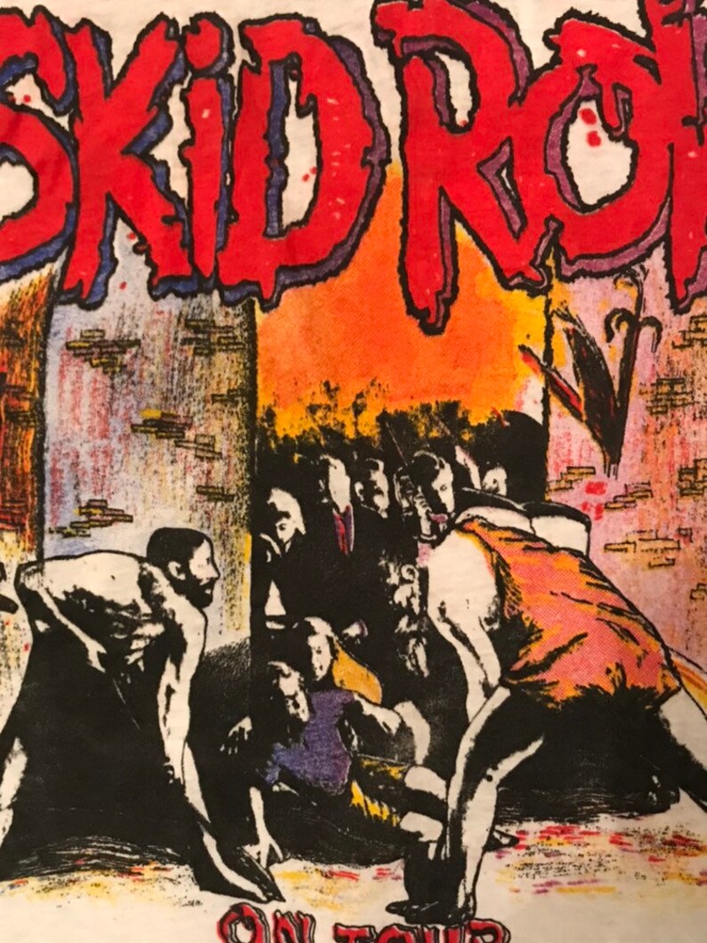 Op de afbeelding: Wit T-shirt met een grafisch ontwerp. De tekst "SKID ROW" is rood met een blauwe omlijning. De afbeelding toont een menigte mensen in een concertsetting, met de woorden "ON TOUR" onderaan.