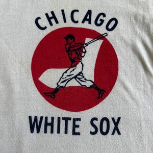 K&ouml;nnte beinhalten: Wei&szlig;es T-Shirt mit einem roten Kreis und einer Grafik eines Baseballspielers. Der Text "CHICAGO WHITE SOX" ist in Marineblau gedruckt.