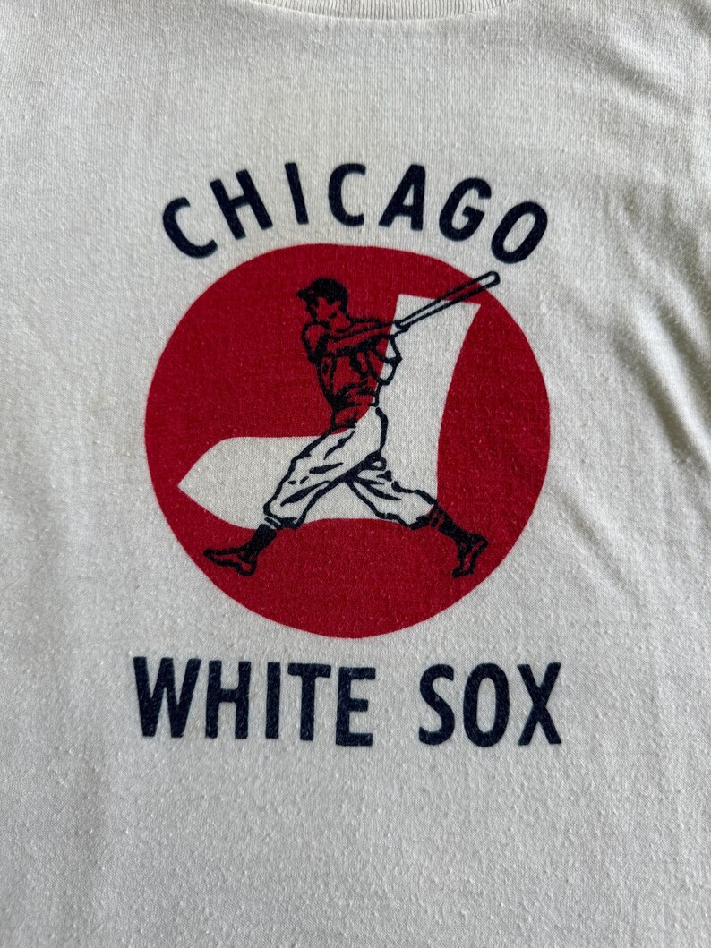 K&ouml;nnte beinhalten: Wei&szlig;es T-Shirt mit einem roten Kreis und einer Grafik eines Baseballspielers. Der Text "CHICAGO WHITE SOX" ist in Marineblau gedruckt.