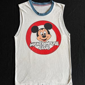 Puede incluir: Camiseta blanca sin mangas con un círculo rojo y la cara de Mickey Mouse en el centro. El texto "Mickey Mouse Club" está escrito en un círculo rojo alrededor de la cara de Mickey Mouse.