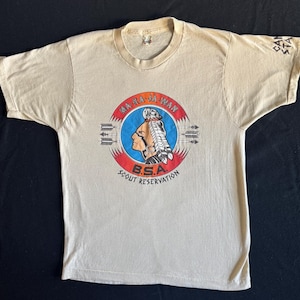 May include: Beige t-shirt featuring a circular graphic with a Native American profile, arrows, and text that reads "MA-RA-JA-WAN B.S.A. SCOUT RESERVATION." The sleeve has text that reads "SA KA".