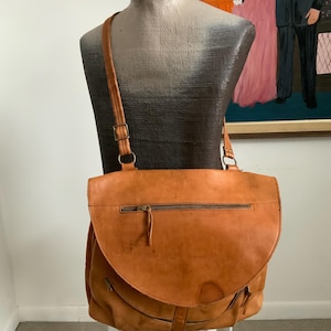 Könnte beinhalten: Eine braune Leder-Umhängetasche mit gebogenem Überschlag und einem langen, verstellbaren Schultergurt. Die Tasche hat ein Hauptfach mit Reißverschluss und zusätzliche Reißverschlusstaschen. Die Tasche wird auf einer Schaufensterpuppe präsentiert.