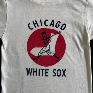 K&ouml;nnte beinhalten: Wei&szlig;es T-Shirt mit einem roten Kreis und einer Grafik eines Baseballspielers. Der Kreis tr&auml;gt den Text "CHICAGO" dar&uuml;ber und "WHITE SOX" darunter.