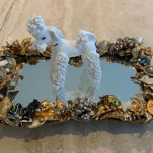 May include: A decorative mirrored tray featuring a white poodle figurine. The tray's frame is embellished with gold, silver, and pearl-like adornments, including various decorative elements in different shapes and sizes.