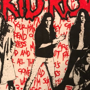 Op de afbeelding: Wit T-shirt met een rood-zwart grafisch ontwerp. Het ontwerp bevat het woord "KIDROCK" in het rood, met zwarte contouren, en verschillende figuren in het zwart. De achtergrond heeft rode handgeschreven tekst.