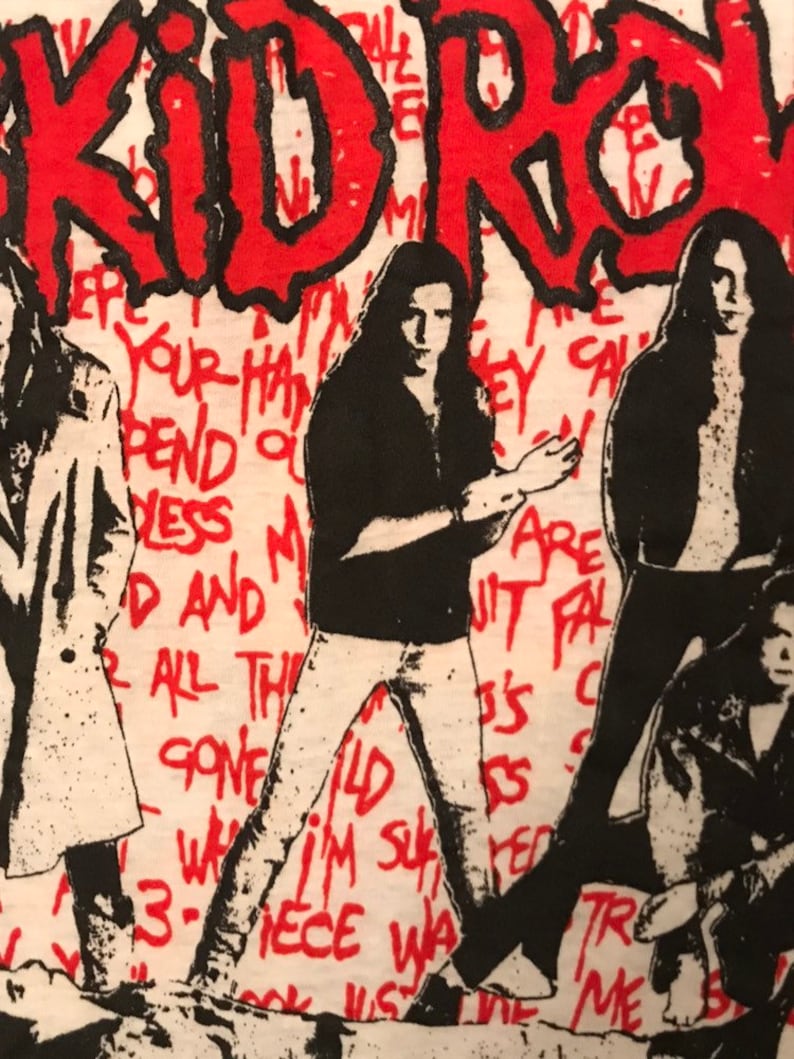 Op de afbeelding: Wit T-shirt met een rood-zwart grafisch ontwerp. Het ontwerp bevat het woord "KIDROCK" in het rood, met zwarte contouren, en verschillende figuren in het zwart. De achtergrond heeft rode handgeschreven tekst.