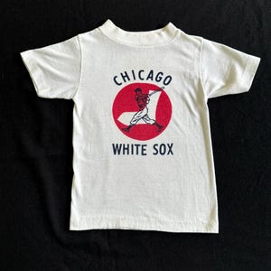 K&ouml;nnte beinhalten: Wei&szlig;es T-Shirt mit einem roten Kreis und einer Grafik eines Baseballspielers. Der Text "CHICAGO WHITE SOX" ist auf das Shirt gedruckt.