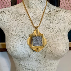 May include: A gold-toned pendant necklace featuring a square, gray center with a raised design. The pendant is set within a decorative gold frame and hangs from a matching gold chain. The necklace is displayed on a mannequin.