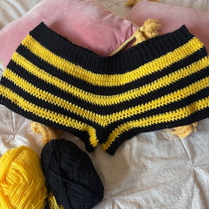 May include: Hand-crocheted shorts in a black and yellow striped pattern, resembling a bee. Two balls of yarn, one yellow and one black, are next to the shorts. The shorts are displayed on a bed with pink pillows.