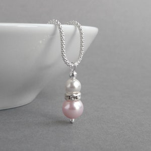 May include: A silver chain necklace with a pink and white pearl pendant. The pendant features a smaller white pearl on top of a larger pink pearl, with a silver band of rhinestones between the two pearls.