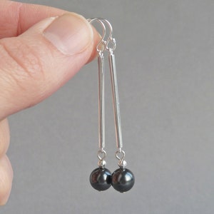 Puede incluir: Pendientes de plata con gotas de perlas negras. Los pendientes tienen una barra larga y delgada de plata con una peque&ntilde;a perla negra en el extremo.
