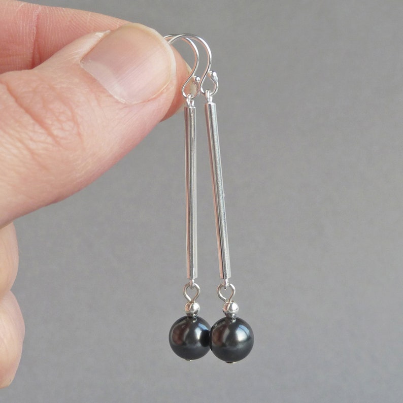 Puede incluir: Pendientes de plata con gotas de perlas negras. Los pendientes tienen una barra larga y delgada de plata con una peque&ntilde;a perla negra en el extremo.