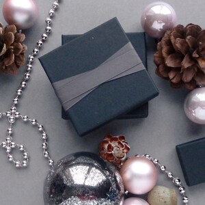 May include: A dark gray gift box with a gray satin ribbon. The box is surrounded by silver beads, pine cones, and silver and pink ornaments.