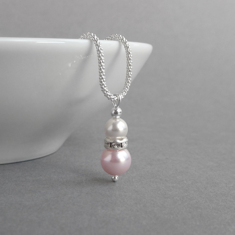 May include: A silver chain necklace with a pink and white pearl pendant. The pendant features a smaller white pearl on top of a larger pink pearl, with a silver band of rhinestones between the two pearls.