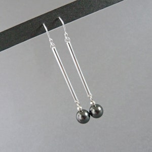 Puede incluir: Un par de pendientes de plata con detalles de perlas negras. Los pendientes tienen una barra larga y delgada de plata con una perla negra en la parte inferior.