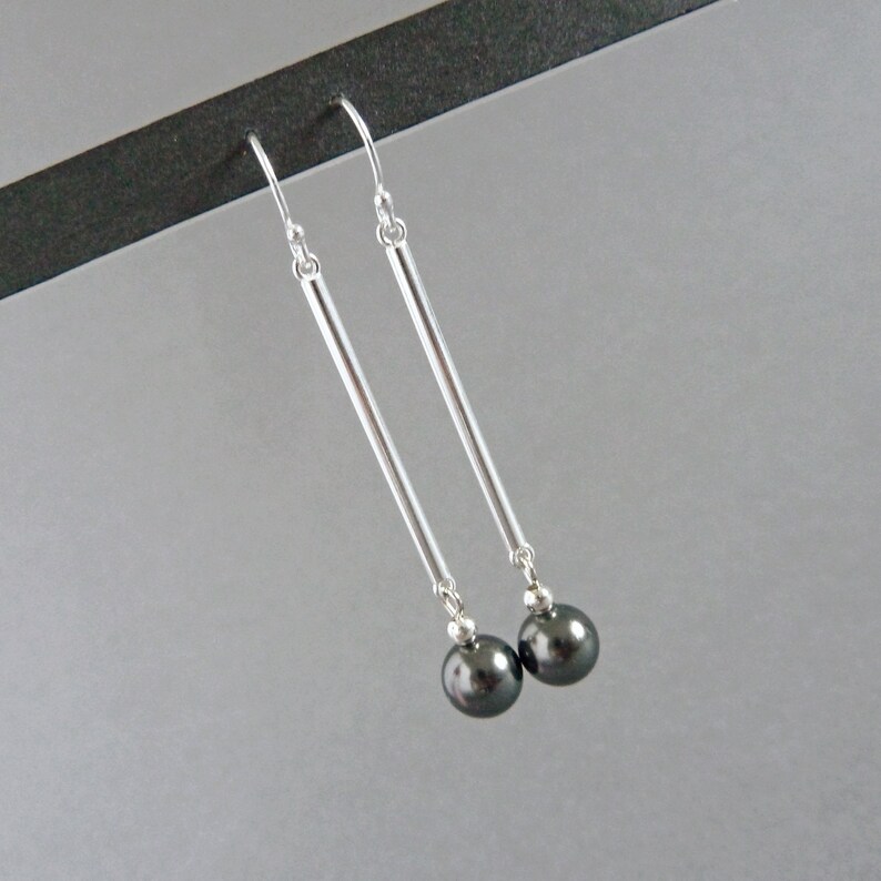 Puede incluir: Un par de pendientes de plata con detalles de perlas negras. Los pendientes tienen una barra larga y delgada de plata con una perla negra en la parte inferior.