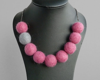 Könnte beinhalten: Eine Halskette aus rosa und grauen Filzkugeln. Die Halskette befindet sich auf einer schwarzen Puppe.