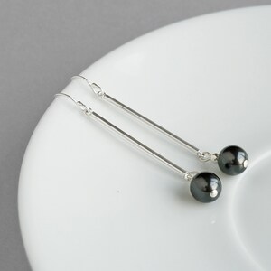 Puede incluir: Un par de pendientes de plata con detalles de perlas negras. Los pendientes tienen una barra larga y delgada de plata con una perla negra en el extremo.