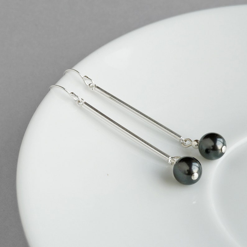 Puede incluir: Un par de pendientes de plata con detalles de perlas negras. Los pendientes tienen una barra larga y delgada de plata con una perla negra en el extremo.