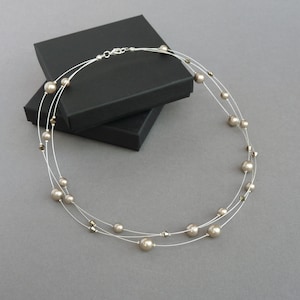 May include: A multi-strand necklace with beige pearls and silver accents. The necklace is on a grey surface and is displayed in a black box.