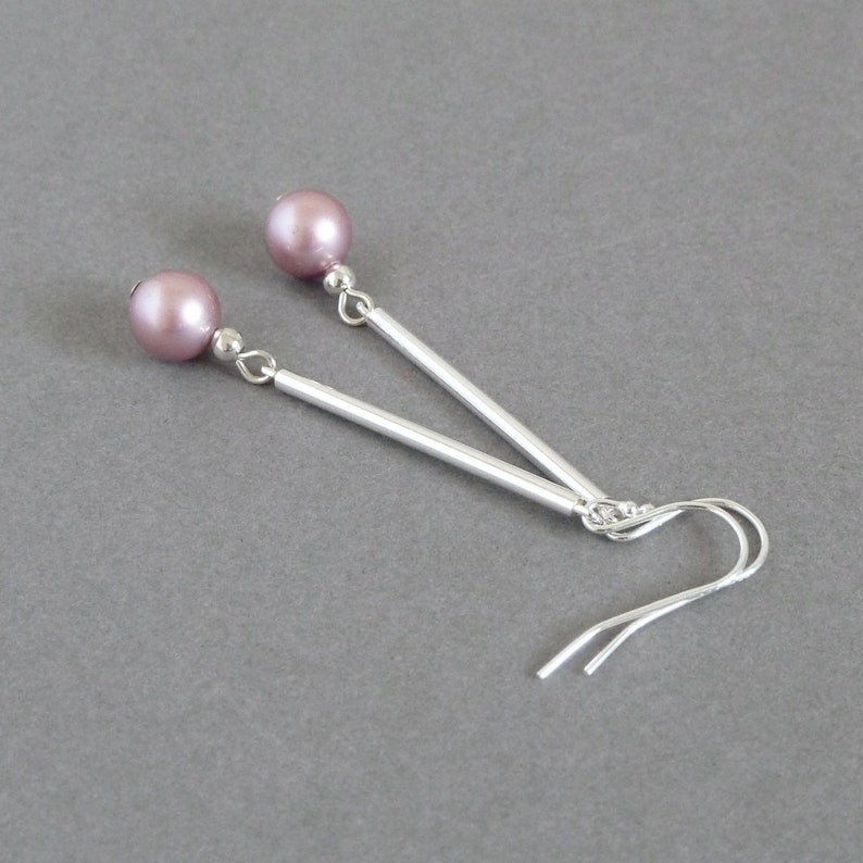 May include: A pair of silver earrings with a mauve pearl on each earring. The earrings have a long, straight silver bar and a small silver hook.