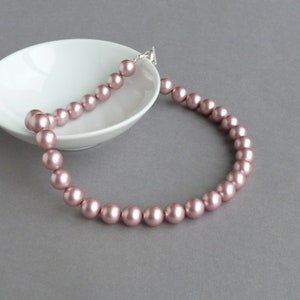 May include: A pink pearl necklace with a silver clasp. The necklace is composed of round, lustrous pearls of varying sizes. The necklace is resting on a gray surface next to a white bowl.