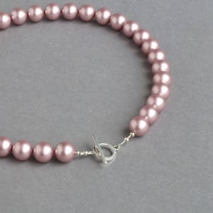 May include: A necklace featuring a strand of pink faux pearls. The necklace is secured with a silver-colored toggle clasp. The pearls are a soft, muted pink color and are evenly spaced along the strand.