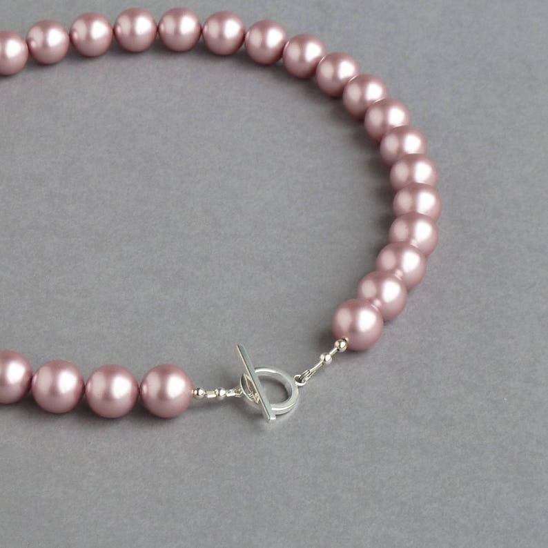 May include: A necklace featuring a strand of pink faux pearls. The necklace is secured with a silver-colored toggle clasp. The pearls are a soft, muted pink color and are evenly spaced along the strand.