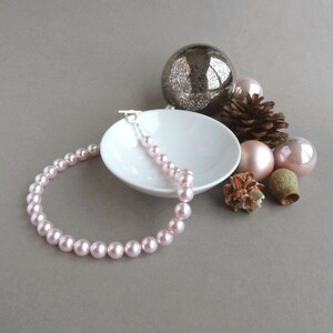 May include: A pink pearl necklace with a silver clasp. The necklace is displayed on a white saucer with decorative ornaments.