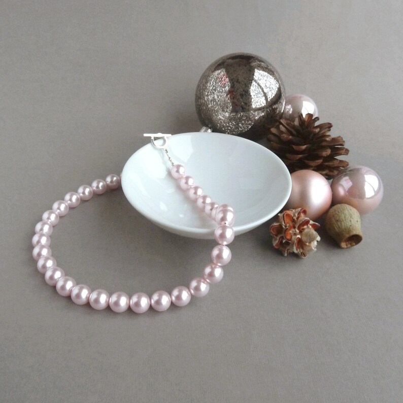 May include: A pink pearl necklace with a silver clasp. The necklace is displayed on a white saucer with decorative ornaments.