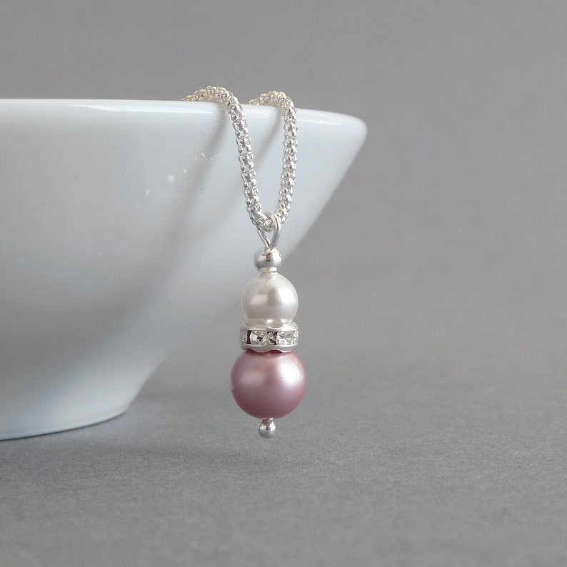 May include: A silver chain necklace with a two-tiered pendant featuring a white pearl and a pink pearl with a clear rhinestone band.