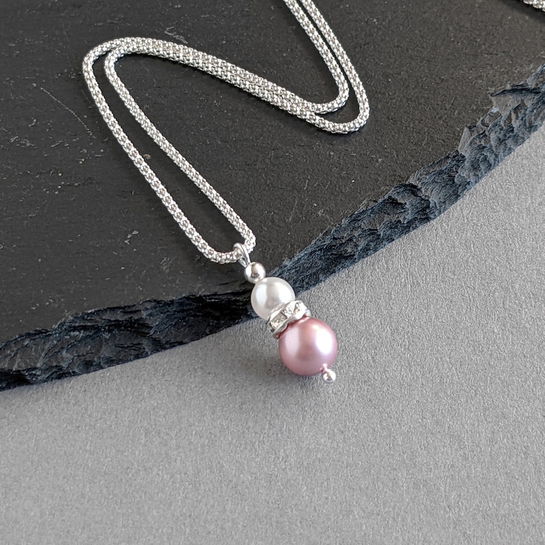 May include: A silver chain necklace with a pink and white pearl pendant. The pendant features a smaller white pearl topped with a clear rhinestone.